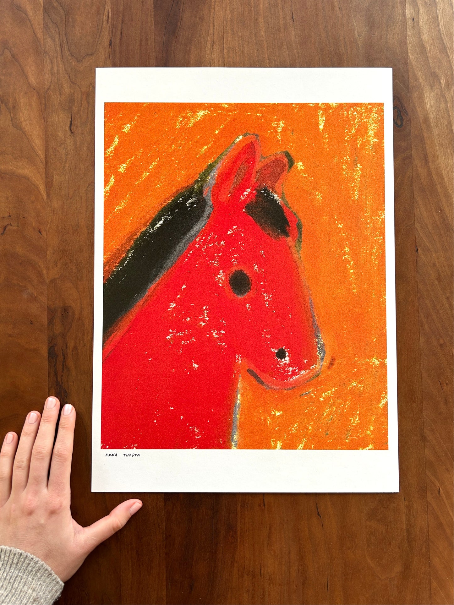 Abstract art print of a red horse head on an orange background, held by a hand against a wooden surface.